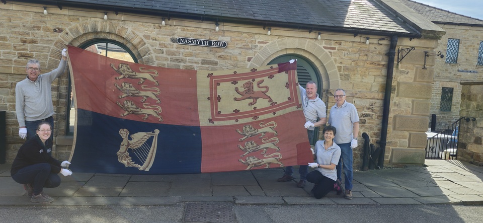 Six people posing outside a stone building, holding and kneeling around a large, historic flag featuring three lions, a single lion, and a harp, each in separate quadrants. The group is smiling and wearing gloves.